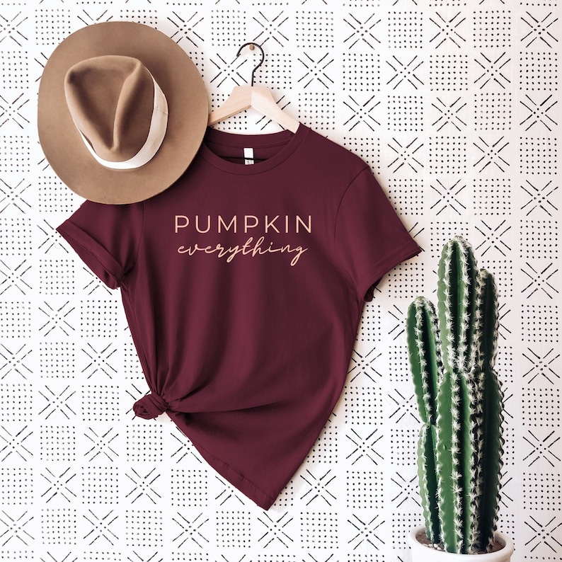 Puede incluir: Una camiseta de color granate con el texto "PUMPKIN everything" en color rosa claro. La camiseta est&aacute; colgada de una percha sobre un fondo blanco con un patr&oacute;n geom&eacute;trico negro.