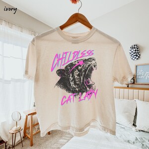 May include: A light beige cropped t-shirt with a graphic print of a roaring tiger with the text "Childless Cat Lady" in pink and blue.