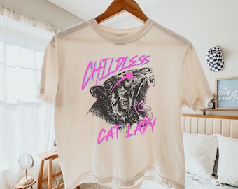 May include: A light beige cropped t-shirt with a graphic print of a roaring tiger with the text "Childless Cat Lady" in pink and blue.