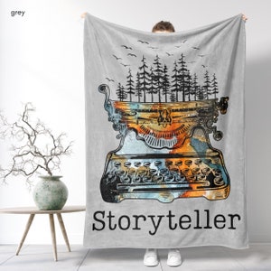 Könnte beinhalten: Eine graue Fleece-Decke mit einer Illustration einer alten Schreibmaschine. Die Schreibmaschine ist von einer Aquarell-Waldlandschaft umgeben. Der Text "Storyteller" ist unter der Schreibmaschine gedruckt.