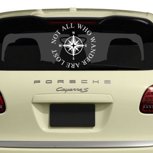 May include: White vinyl car decal featuring a compass rose design within a circular frame. The text reads "NOT ALL WHO WANDER ARE LOST." The decal is applied to the rear window of a cream-colored Porsche Cayenne S.