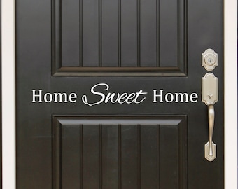 May include: A black front door with a white "Home Sweet Home" vinyl decal. The door has a silver door handle and a silver deadbolt lock.