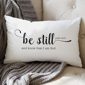 May include: White throw pillow with black text that reads "be still Psalm 46:10 and know that I am God".