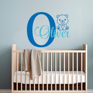 May include: A blue and light blue personalized wall decal with the letter O and the name Oliver. A blue teddy bear with a bow tie is sitting on top of the letter O. The decal is displayed on a light blue wall above a white crib.