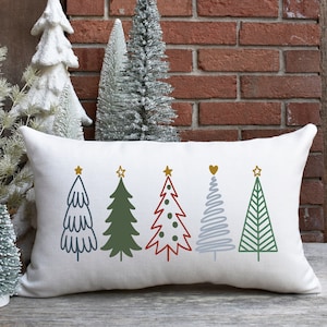 May include: A white pillow with five different colored Christmas trees embroidered on it. The trees are green, red, blue, and gray. Each tree has a gold star on top.