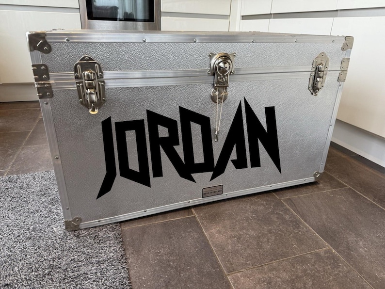 Pu&ograve; includere: Un baule in metallo argentato con il nome "JORDAN" in grandi lettere nere angolari sulla parte anteriore. Il baule ha ferramenta in metallo, tra cui chiusure e protezioni angolari. Il baule &egrave; appoggiato su un pavimento.