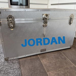 Pu&ograve; includere: Un baule in metallo argentato con il nome "JORDAN" in lettere blu. Il baule ha protezioni angolari in metallo, chiusure e una superficie strutturata. &Egrave; appoggiato su un tappeto grigio e un pavimento in piastrelle marroni.