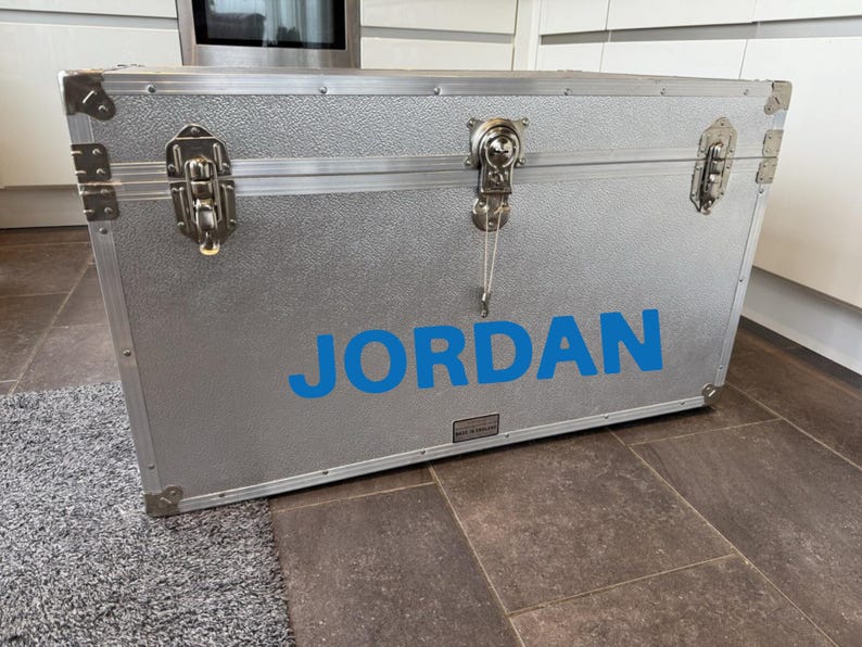 Pu&ograve; includere: Un baule in metallo argentato con il nome "JORDAN" in lettere blu. Il baule ha protezioni angolari in metallo, chiusure e una superficie strutturata. &Egrave; appoggiato su un tappeto grigio e un pavimento in piastrelle marroni.