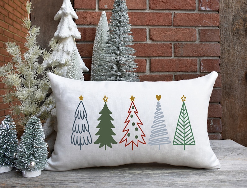 May include: A white pillow with five different colored Christmas trees embroidered on it. The trees are green, red, blue, and gray. Each tree has a gold star on top.