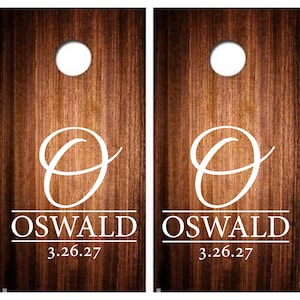 May include: Two wooden cornhole boards with a dark brown finish. Each board features a white letter "O" and the name "OSWALD" in white, along with the date "3.26.27". A white hole is at the top of each board.