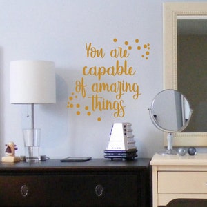 May include: Gold vinyl wall decal with the text "You are capable of amazing things" with small gold dots around the text.