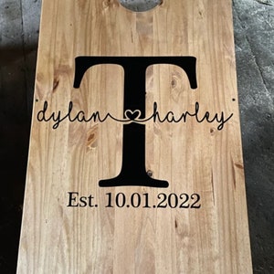 May include: A wooden cornhole board with a large black letter "T" in the center. The names "dylan" and "harley" are written in cursive on either side of the letter, connected by a heart. The date "Est. 10.01.2022" is below.