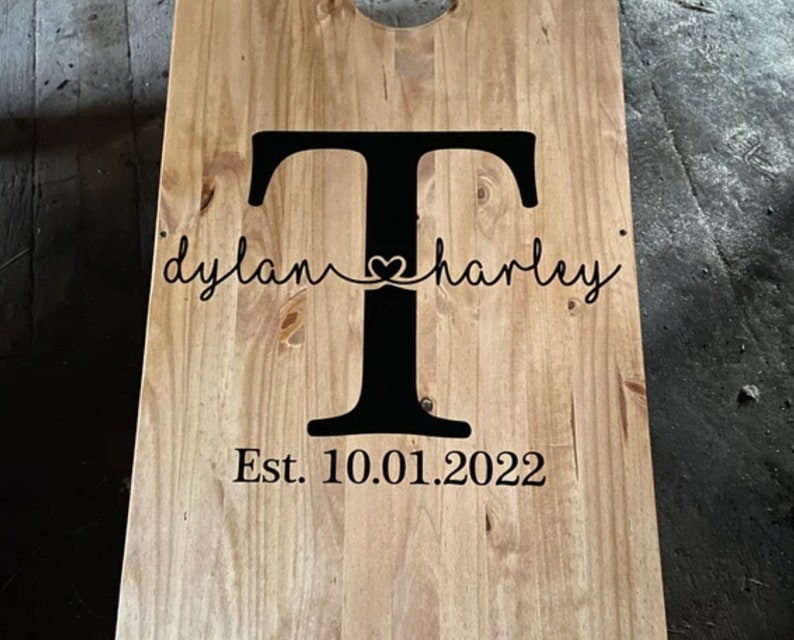 May include: A wooden cornhole board with a large black letter "T" in the center. The names "dylan" and "harley" are written in cursive on either side of the letter, connected by a heart. The date "Est. 10.01.2022" is below.