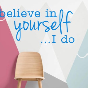 May include: A wooden chair sits in front of a wall with a geometric pattern of pink, gray, and blue triangles. The wall has the text "believe in yourself... I do" written in blue.