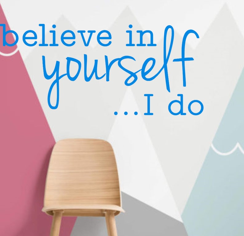 May include: A wooden chair sits in front of a wall with a geometric pattern of pink, gray, and blue triangles. The wall has the text "believe in yourself... I do" written in blue.