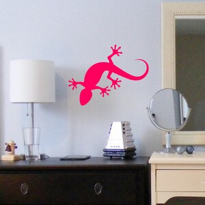 May include: A bright pink gecko silhouette wall decal, a popular home decor item, is shown on a white wall. The decal is positioned above a dark brown dresser with a lamp and a glass of water on top.