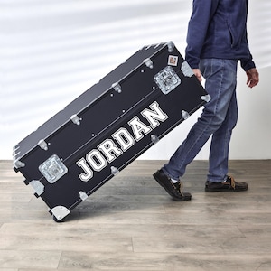 May include: A black trunk with the name "JORDAN" in white block letters. The trunk has silver hardware, corner protectors, and wheels. The trunk is being pulled across a wooden floor.