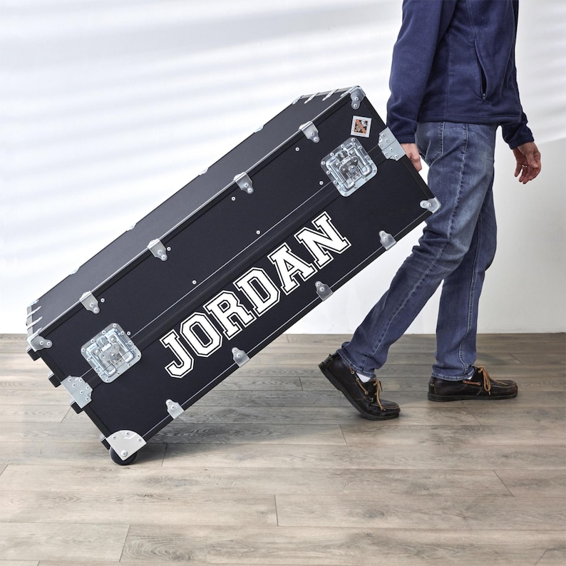 Pu&ograve; includere: Un baule nero con il nome "JORDAN" a lettere bianche. Il baule ha ferramenta argentata, protezioni angolari e ruote. Il baule viene trascinato su un pavimento di legno.