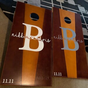 May include: Two cornhole boards with a brown, orange, and tan color scheme. Each board features a large letter "B" with the names "nikki" and "chris" in script, and the date "11.11". The boards have a hole near the top.