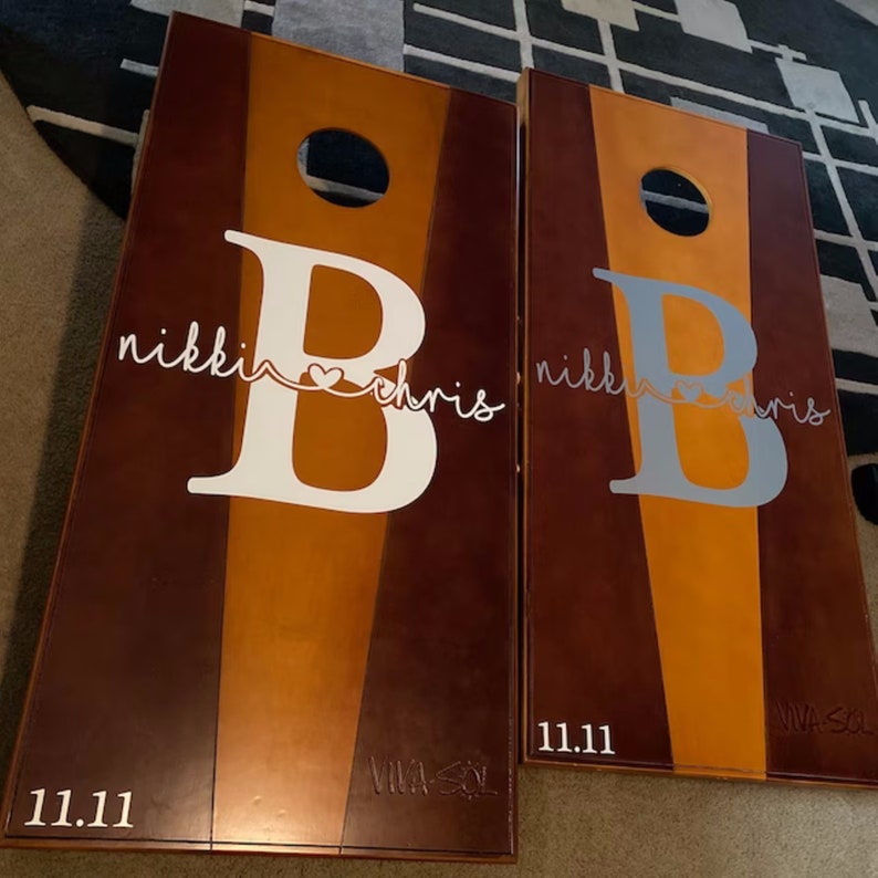 May include: Two cornhole boards with a brown, orange, and tan color scheme. Each board features a large letter "B" with the names "nikki" and "chris" in script, and the date "11.11". The boards have a hole near the top.