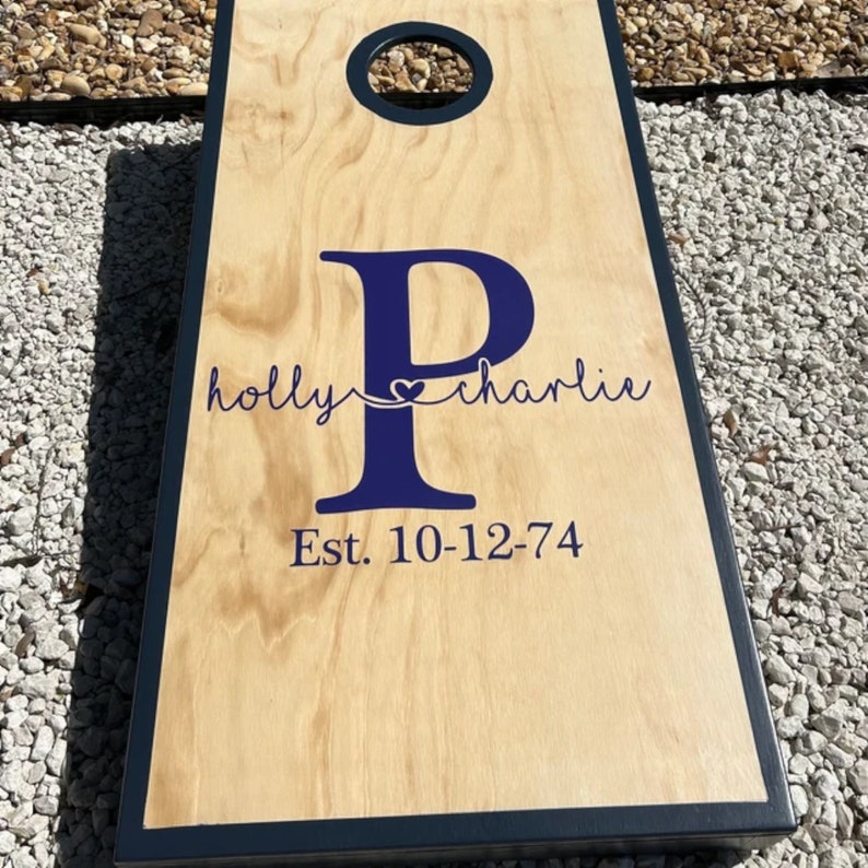 May include: A wooden cornhole board with a navy blue border and a centered hole. The board features a large navy blue letter "P" with the names "holly" and "charlie" and the date "Est. 10-12-74" in navy blue.