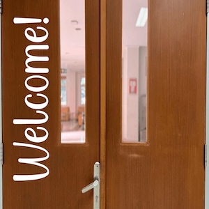 May include: A pair of brown wooden doors with rectangular glass panels. The word "Welcome!" is written vertically in white script on the left door. A silver door handle is visible on the right door.