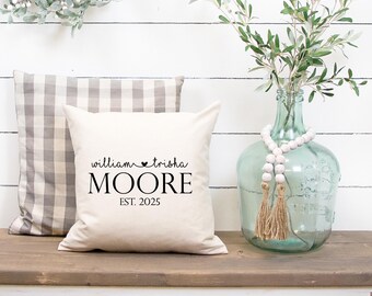 May include: A cream-colored square pillow with the names "William & Trisha Moore" and the year "EST. 2025" printed in black. A gray and white checkered pillow is behind it. A glass vase with greenery and decorative beads is on the right.