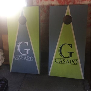 Pode incluir: Duas t&aacute;buas de cornhole de madeira com um design triangular verde e azul. As t&aacute;buas t&ecirc;m um c&iacute;rculo preto no centro do tri&acirc;ngulo e o texto "G GASAPO" em letras brancas.