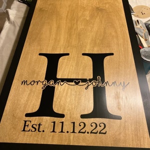 May include: A wooden sign with a light brown surface and a black border. The sign features a large black letter "H" with the names "morgan" and "johnny" in a cursive font, connected by a heart. The date "Est. 11.12.22" is below.
