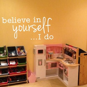 May include: A playroom with a white wall with the words "believe in yourself... I do" written on it. There is a toy kitchen with a pink oven and a white refrigerator. There is a wooden toy shelf with colorful bins filled with toys.