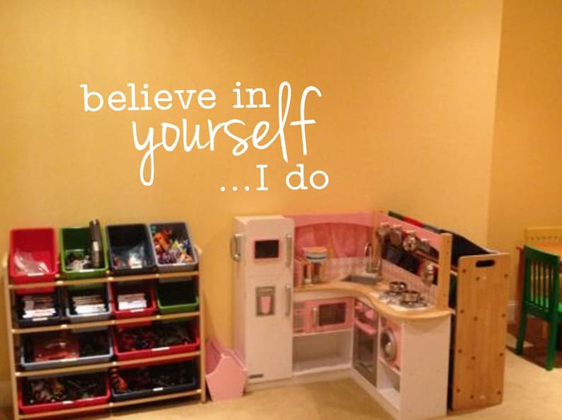 May include: A playroom with a white wall with the words "believe in yourself... I do" written on it. There is a toy kitchen with a pink oven and a white refrigerator. There is a wooden toy shelf with colorful bins filled with toys.