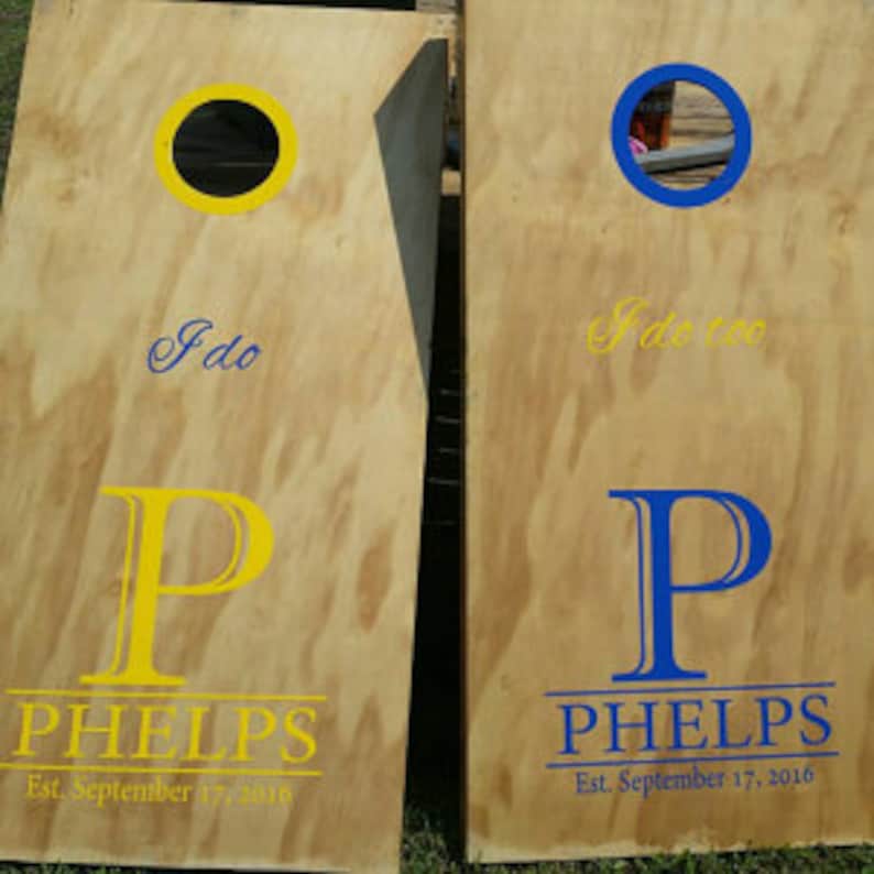 Pode incluir: Dois tabuleiros de cornhole de madeira com um c&iacute;rculo amarelo e azul em cada tabuleiro. Os tabuleiros s&atilde;o personalizados com o texto "I do" e "I do too" e o nome "PHELPS" com a data "Est. September 17, 2016".