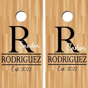 May include: Two wooden cornhole boards with a black and white design. The boards feature the letter "R" with the names "Sandra" and "Carlos" written underneath. The text "RODRIGUEZ" and "Est. 2022" are also included on each board.