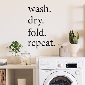 May include: A white washing machine with a black digital display and a white laundry basket with a woven pattern. The wall behind the machine has a black vinyl decal with the text "wash. dry. fold. repeat."