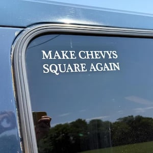 May include: A white decal on a car window with the text "MAKE CHEVYS SQUARE AGAIN". The decal is in a sans-serif font and is adhered to the dark tinted glass of a blue vehicle.