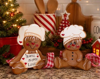 May include: Two brown gingerbread-style figures wearing white chef hats. The left figure wears an apron with the text "BAKE SOME LOVE" and red hearts. Both figures have painted facial features, red cheeks, and are accented with red and white checkered ribbon.