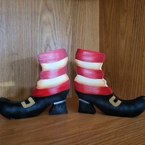 May include: A pair of decorative wooden witch boots. The boots are black with red and white striped tops and gold-colored buckles. They are displayed on a wooden shelf.