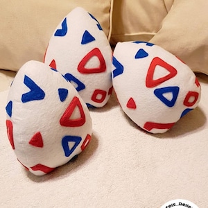 May include: Three white plush egg-shaped pillows with red and blue geometric patterns. The pillows are on a white surface.