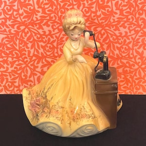 May include: A vintage porcelain figurine of a woman in a yellow dress, holding a black telephone receiver. The figurine has a floral design on the dress and is set against an orange floral background.