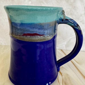 May include: A handmade ceramic mug with a blue handle and a cylindrical body. The mug's lower portion is a deep blue, transitioning to a layered design of blue, gray, and teal at the top. The mug is suitable for hot or cold beverages.