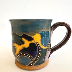 May include: A handmade ceramic mug with a dark teal glaze and a brown handle. The mug features a colorful abstract design with yellow, blue, purple, and black shapes, accented with white dots. The base is a natural tan color.