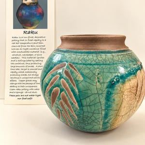 May include: A round ceramic vase with a wide opening and a matte brown rim. The body features a turquoise crackle glaze with a prominent brown leaf-like pattern. Beside it is a card titled Raku with descriptive text about the pottery process.