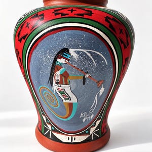 May include: A terracotta-colored ceramic vase featuring a hand-painted scene of a figure playing a flute against a blue speckled background. The vase is decorated with red, green, and black geometric patterns and includes the signature A. Pino 94.