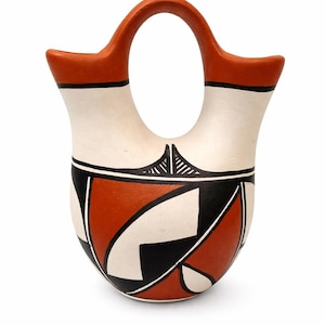 May include: A handcrafted, ceramic water vessel with a distinctive handle. The vessel features a cream-colored body with geometric designs in black and burnt orange. The handle and rim are painted burnt orange with a black accent.