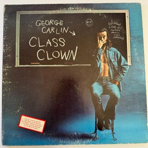 May include: Album cover for George Carlin's "Class Clown." The cover features a chalkboard with the title and artist's name. A man in a denim jacket and jeans is seated on a stool. The album cover has a blue background.