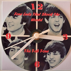 May include: A Beatles themed clock with four images of the band members. The clock face is black with white numbers and hands. The text "Four lads that shook the world" and "The Fab Four" are printed in red.