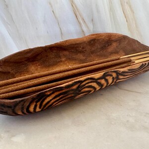 May include: A handcrafted, oblong wooden bowl with a dark brown finish and decorative black and brown wood grain. The bowl contains several brown incense sticks with gold tips, perfect for aromatherapy or meditation.