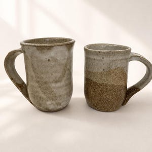 May include: Two handmade ceramic mugs with a rustic, speckled finish. The larger mug on the left features a light grey, mottled glaze. The smaller mug on the right displays a two-tone design with a light grey upper section and a textured, sandy brown base.