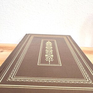 May include: A brown leather-bound book with gold detailing. The cover has a gold rectangular frame with a central floral design. The edges are also trimmed with gold, giving it a classic and elegant appearance.