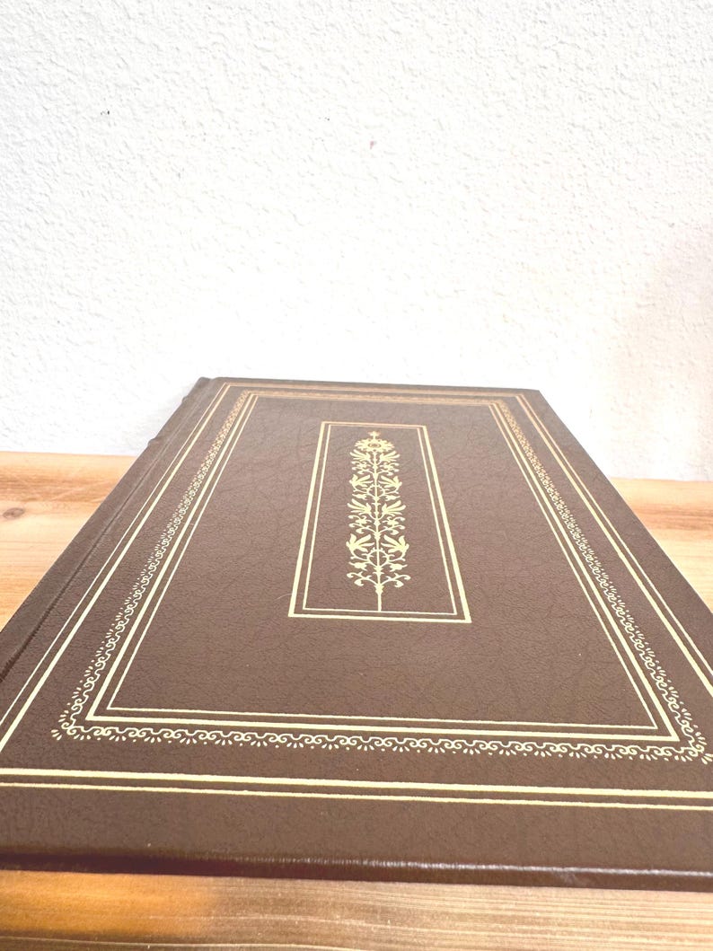 May include: A brown leather-bound book with gold detailing. The cover has a gold rectangular frame with a central floral design. The edges are also trimmed with gold, giving it a classic and elegant appearance.
